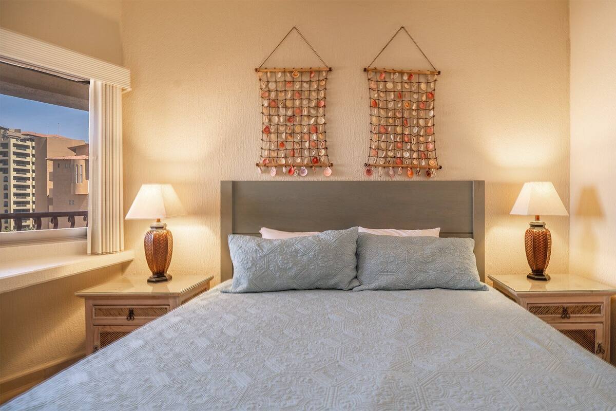 Une chambre d’hôtel avec un grand lit, deux tables de chevet avec des lampes, une fenêtre donnant sur des immeubles et un élément décoratif mural au-dessus du lit.