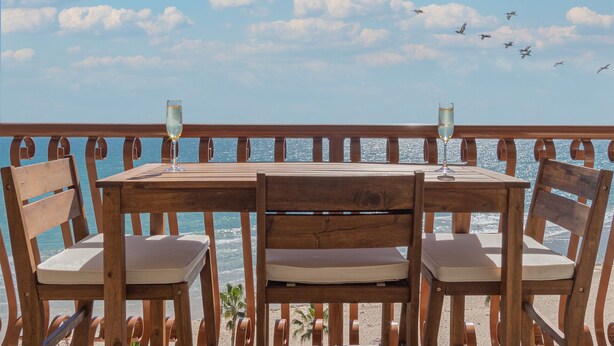 Une table en bois, deux chaises et deux coupes de champagne, avec vue sur une plage bordée d’une balustrade et de palmiers.