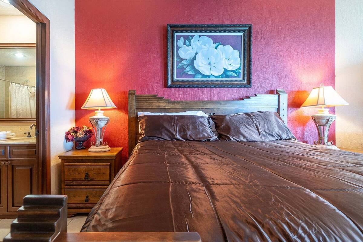 Une chambre d’hôtel avec un grand lit, deux lampes de chevet, une table de chevet en bois avec une lampe, un miroir et une œuvre d’art florale encadrée accrochée au mur.