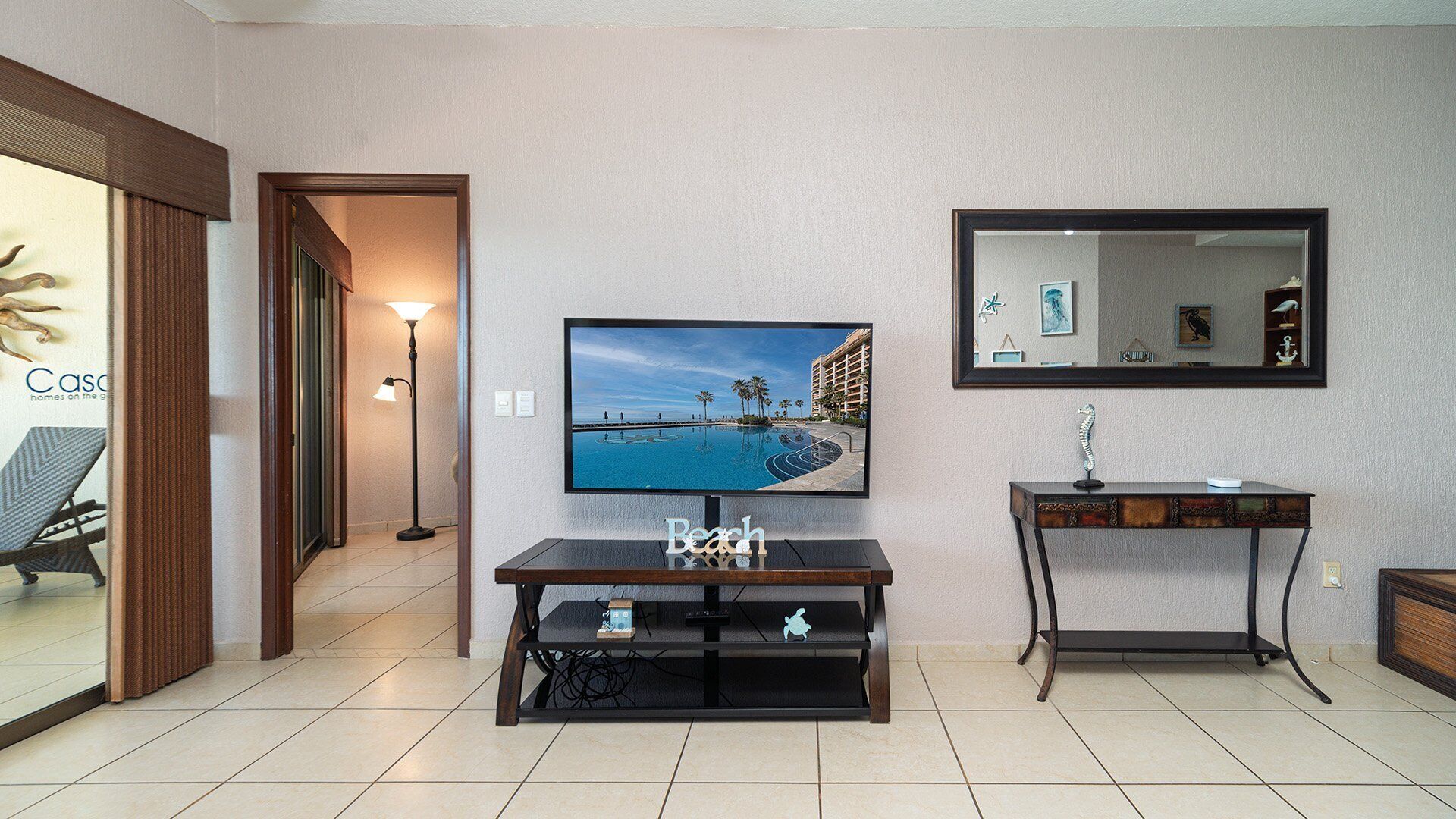 A hotel room with a large flat-screen TV mounted on the wall, a small wooden console table, a mirror, and a view of a pool and building.