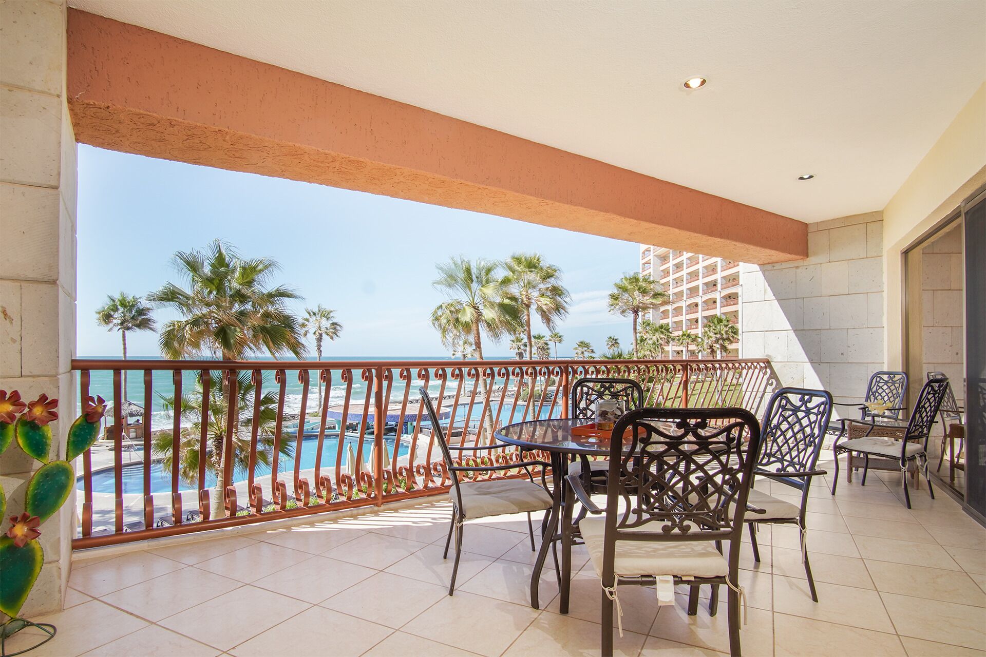 A balcony with a view of a pool and palm trees, furnished with metal chairs and a table.