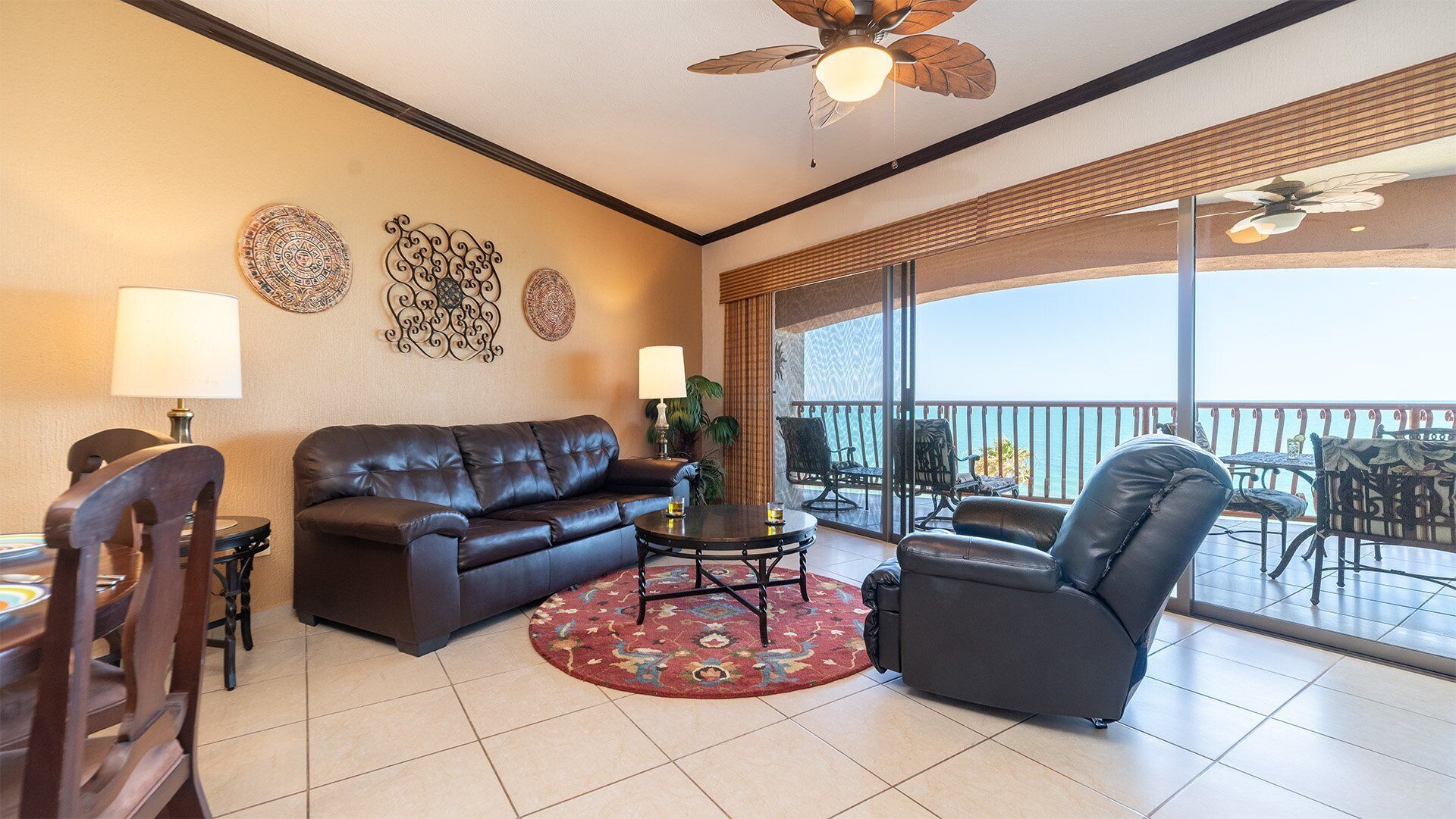 A spacious living room with a dark leather sofa, a round coffee table, and a black armchair. There are decorative wall art pieces and a large window offering a view of the ocean.