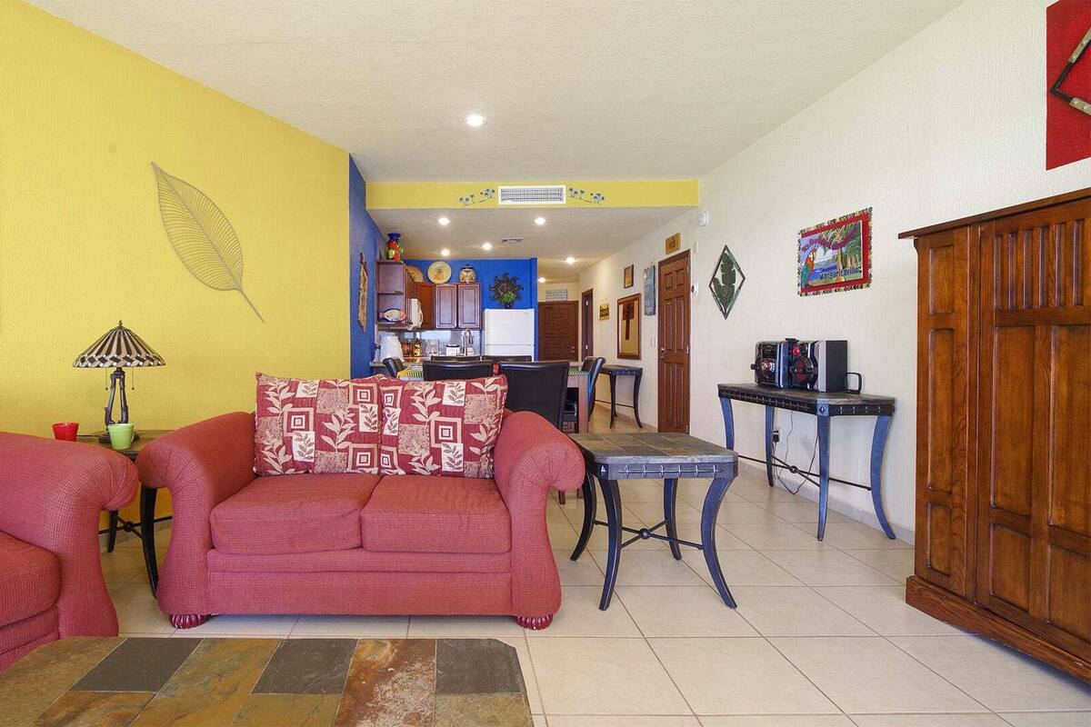 A living room with two red sofas, a patterned throw pillow, a small table with a lamp, and a kitchen area in the background.