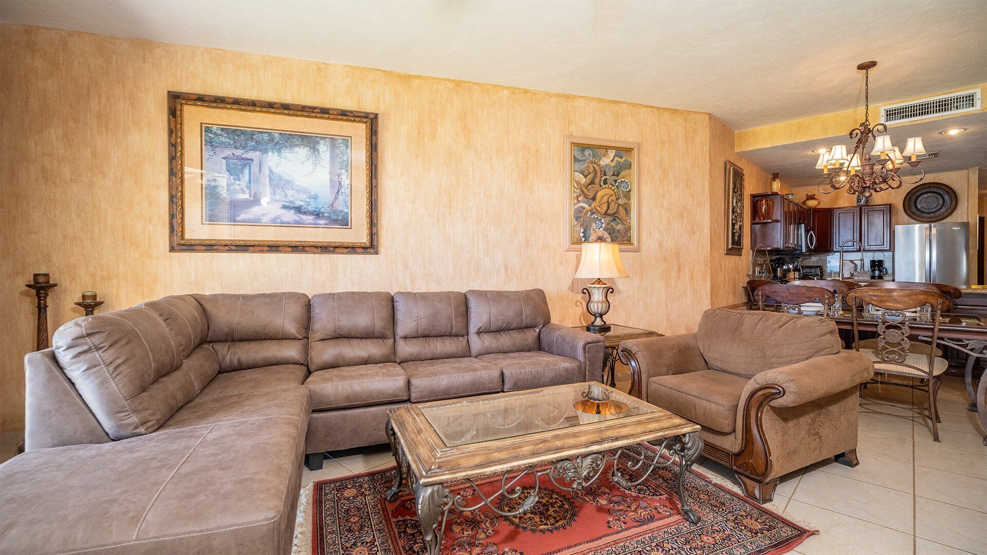 Ein Wohnzimmer mit einem großen L-förmigen Sofa, einem kleineren Sessel, einem Holztisch und einem gemusterten Teppich. An den Wänden hängen zwei Bilder, eine Chandelieber und im Hintergrund ist eine Küchenzeile erkennbar.