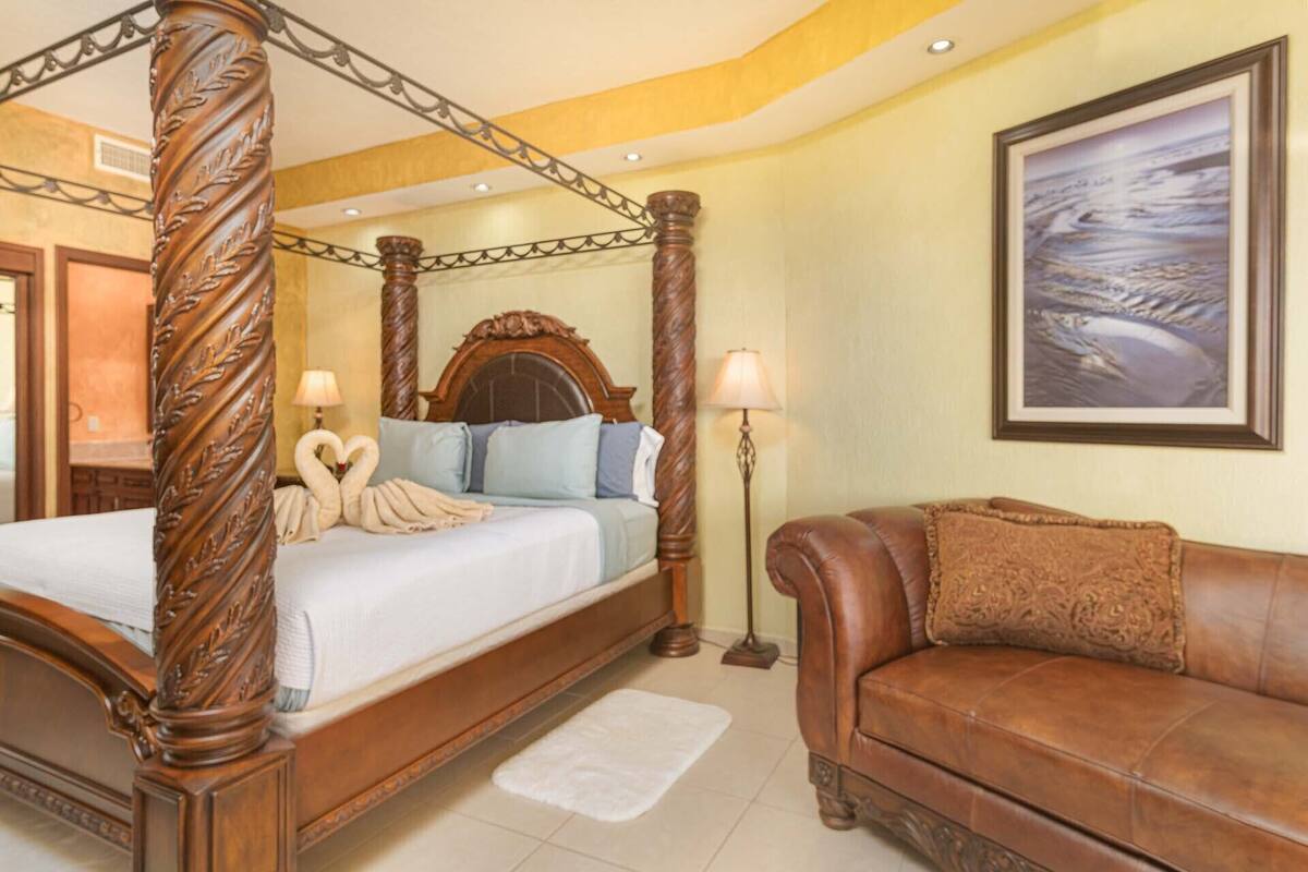 A four-poster bed with a heart-shaped towel arrangement, a brown leather sofa, and a framed landscape painting on the wall.