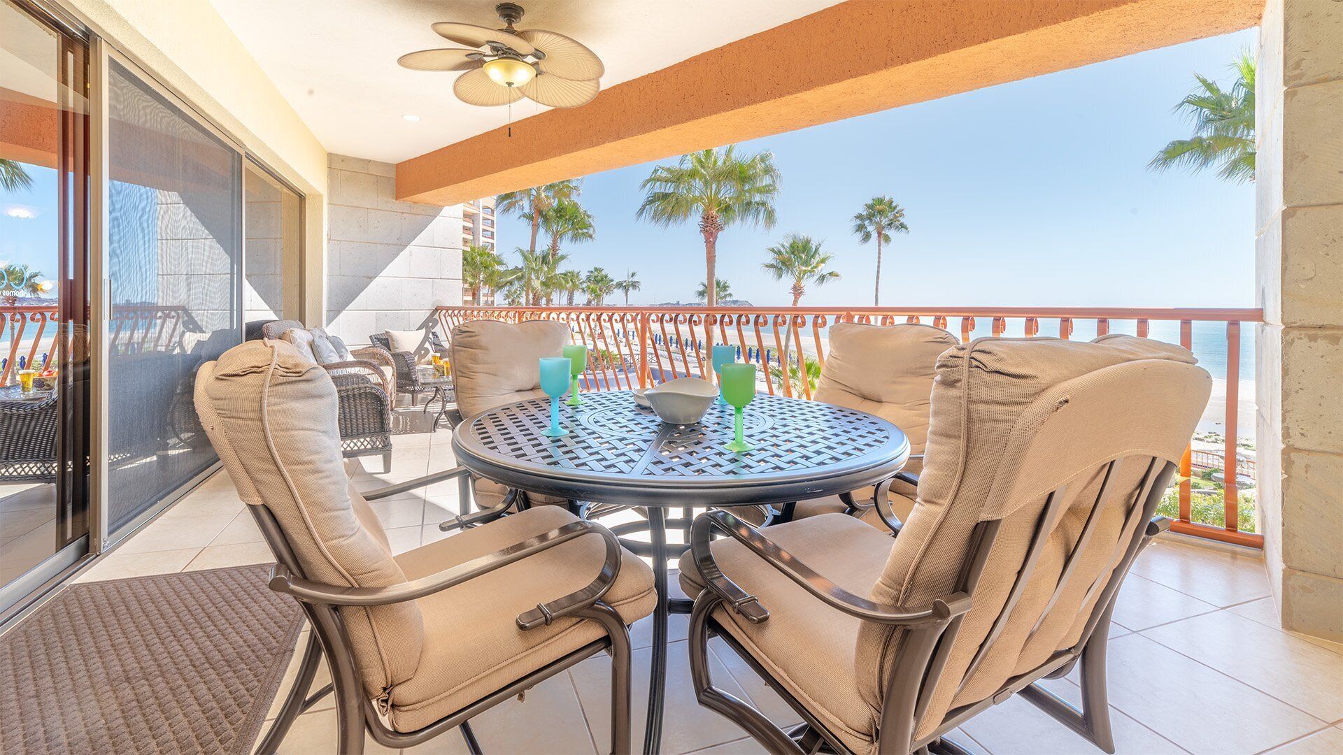 Ein Balkon mit rundem Tisch und Stühlen, einem Deckenventilator und Blick auf Palmen und den Strand.