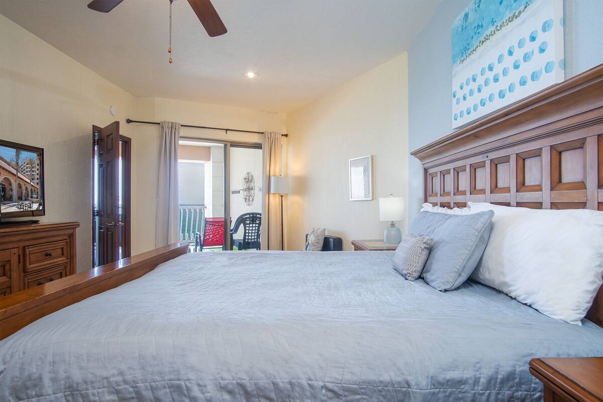 A hotel room with a large bed, a television on a wooden dresser, a balcony with a view, and a ceiling fan.