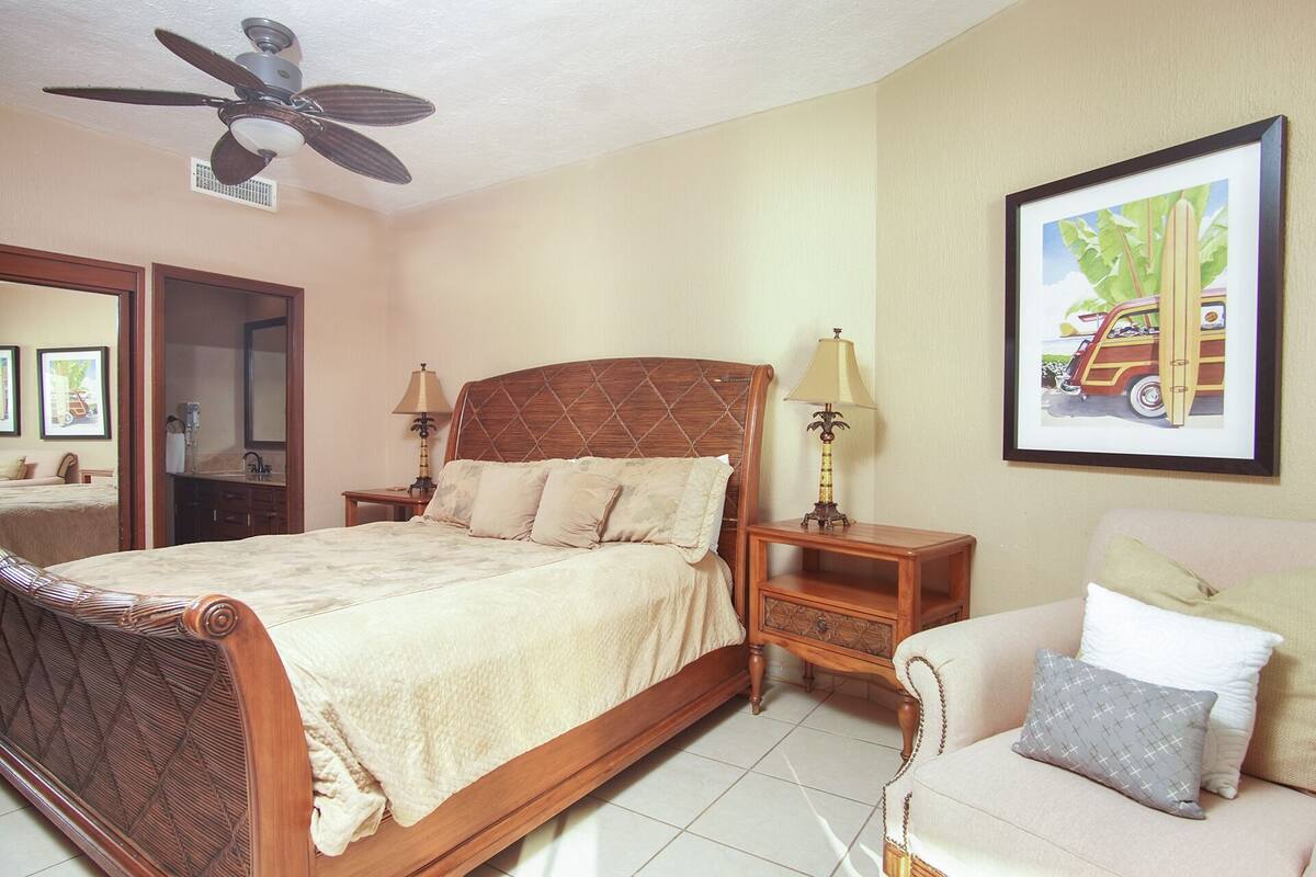 A hotel room with a wooden bed, a ceiling fan, a nightstand with a lamp, a framed picture on the wall, and a sofa with pillows.