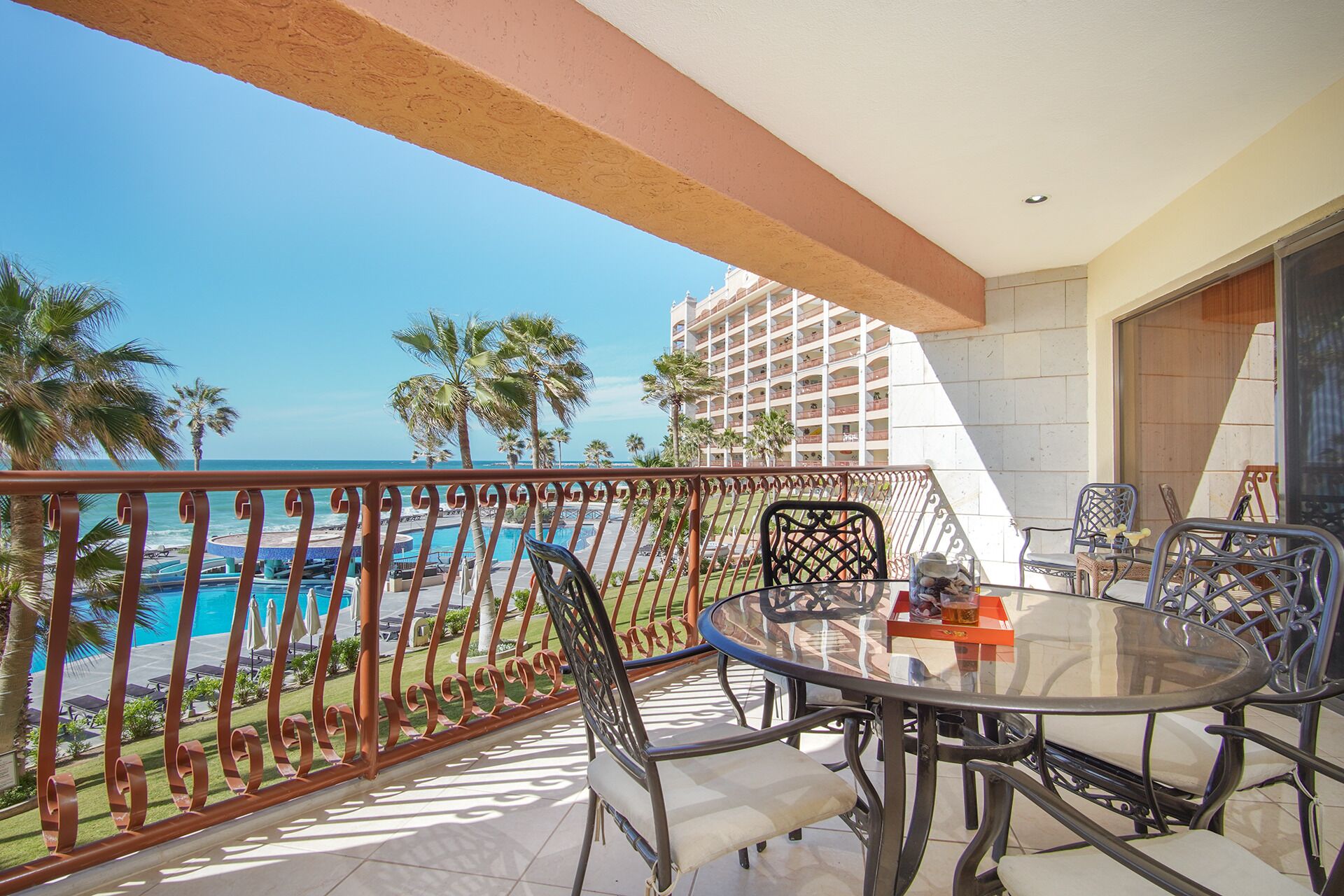 A balcony with a glass table and chairs, overlooking a pool and palm trees.
