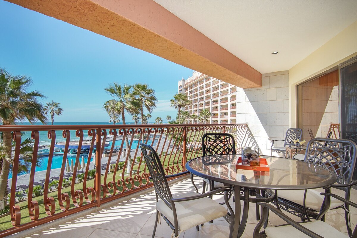 A balcony with a glass table and chairs, overlooking a pool and palm trees.