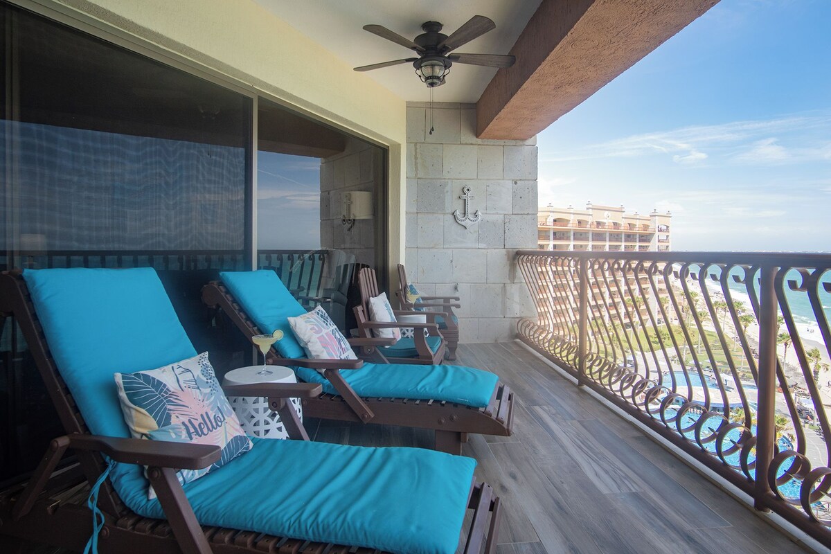 A balcony with wooden lounge chairs, a ceiling fan, and a view of a beach and hotel buildings.