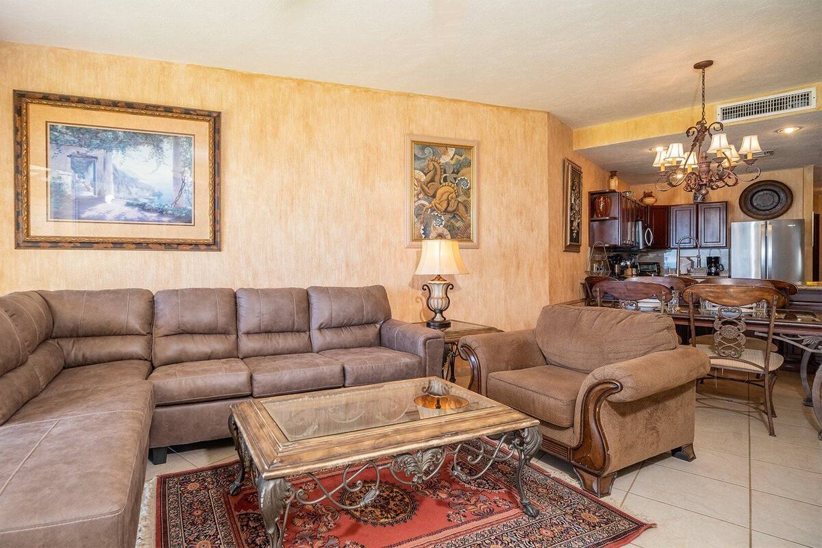 Ein Wohnzimmer mit einem großen L-förmigen Sofa, einem kleineren Sessel, einem Holztisch und einem gemusterten Teppich. An den Wänden hängen zwei Bilder, eine Chandelieber und im Hintergrund ist eine Küchenzeile erkennbar.