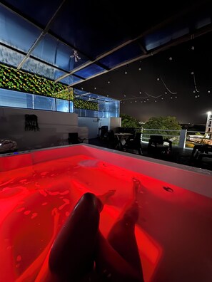 Uma área de piscina no terraço, com um elemento aquático iluminado em vermelho, mobiliário de exterior e vista para a cidade à noite.
