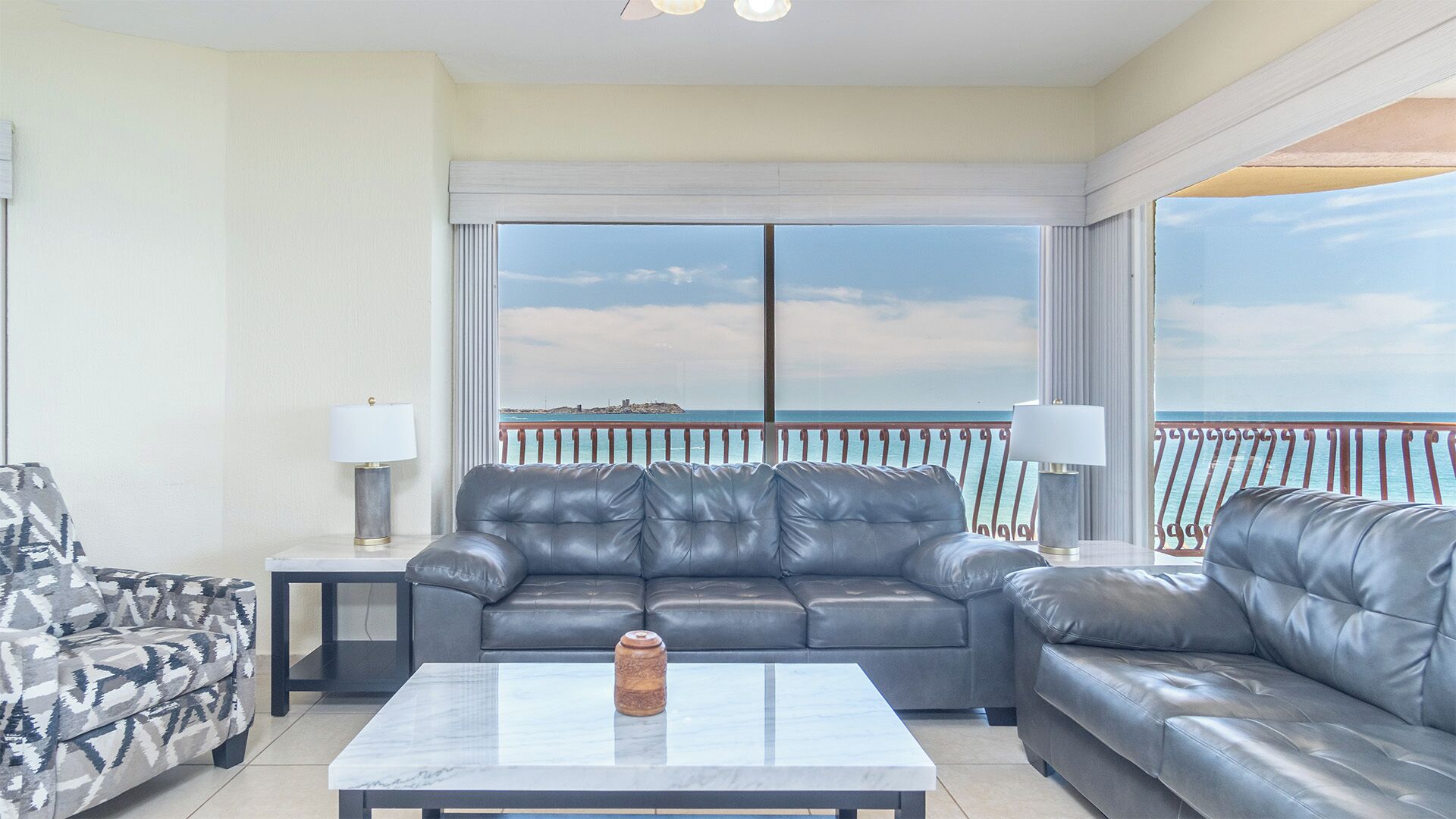 A spacious balcony with a view of the sea, a black leather sofa, a patterned armchair, and a marble coffee table.