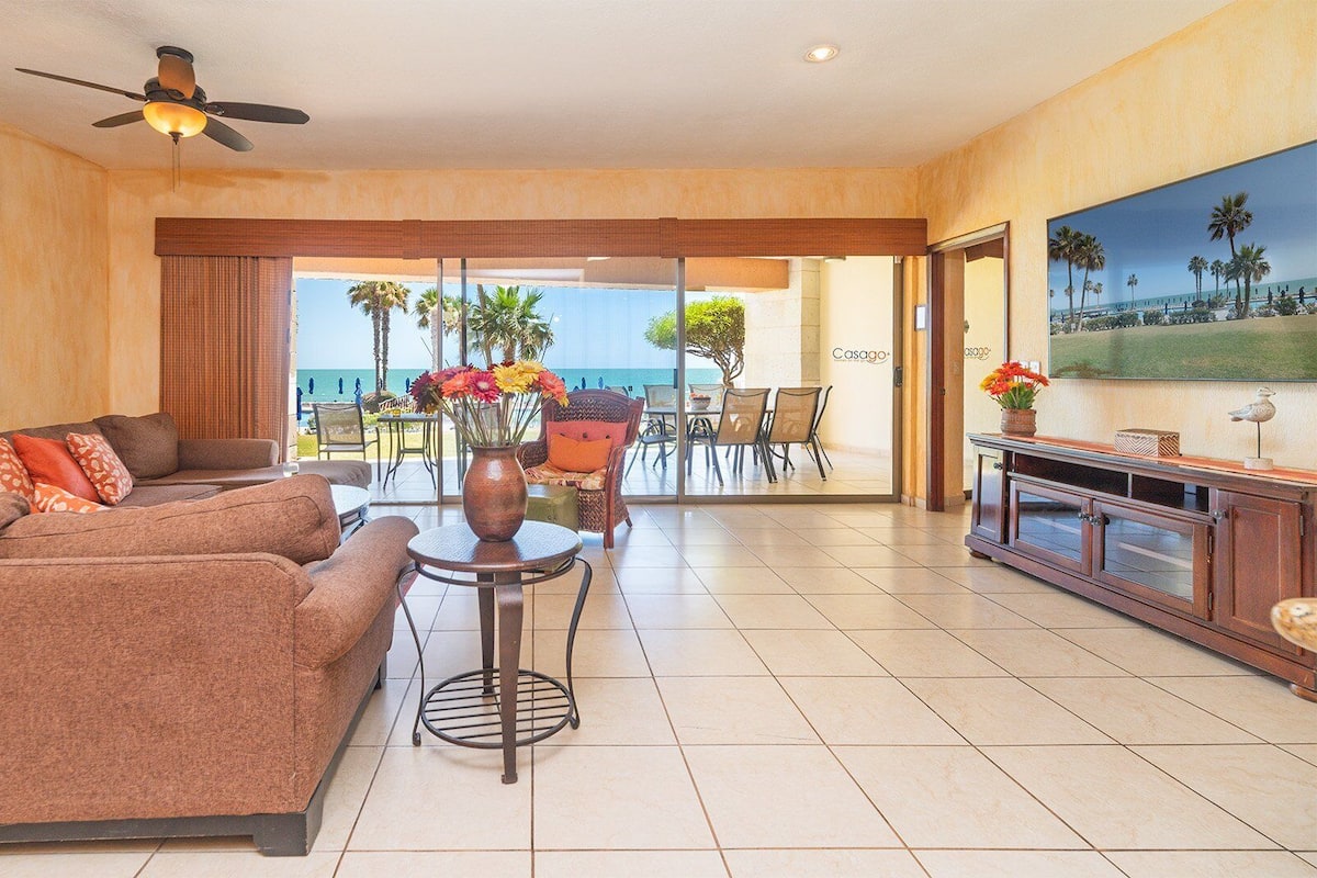 A hotel room with a sofa, a small table with a vase of flowers, a ceiling fan, and a large window overlooking the beach.