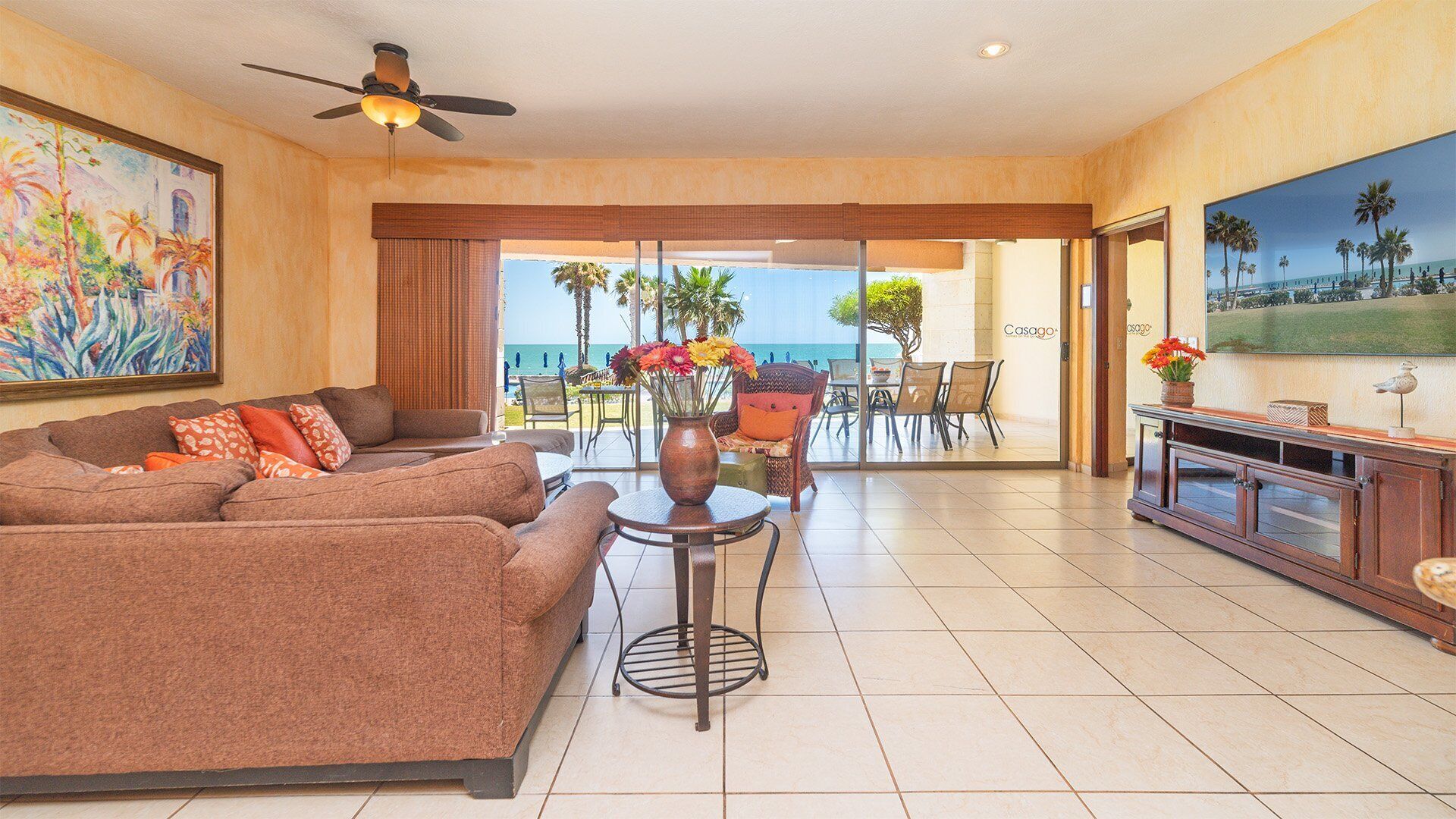 Ein Hotelzimmer mit einer Couch, einem kleinen Tisch mit Blumenvase, einem Deckenventilator und einem großen Fenster mit Blick auf den Strand.