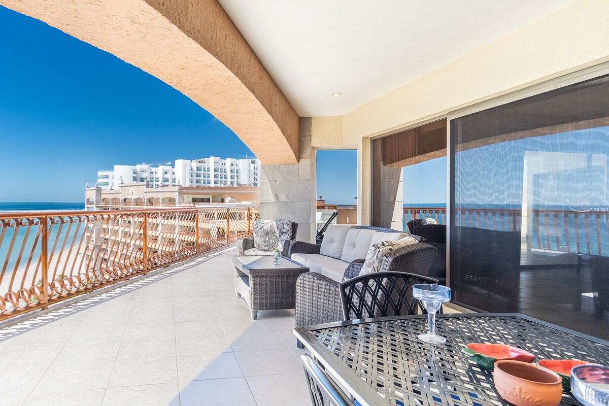 A balcony with a view of the sea and buildings, furnished with wicker chairs and a table with a glass.