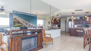 Ein geräumiges Wohnzimmer mit großem Flachbildfernseher, Holzmöbeln, einer Küche mit dunklen Schränken und Meerblick durch eine Glaswand.