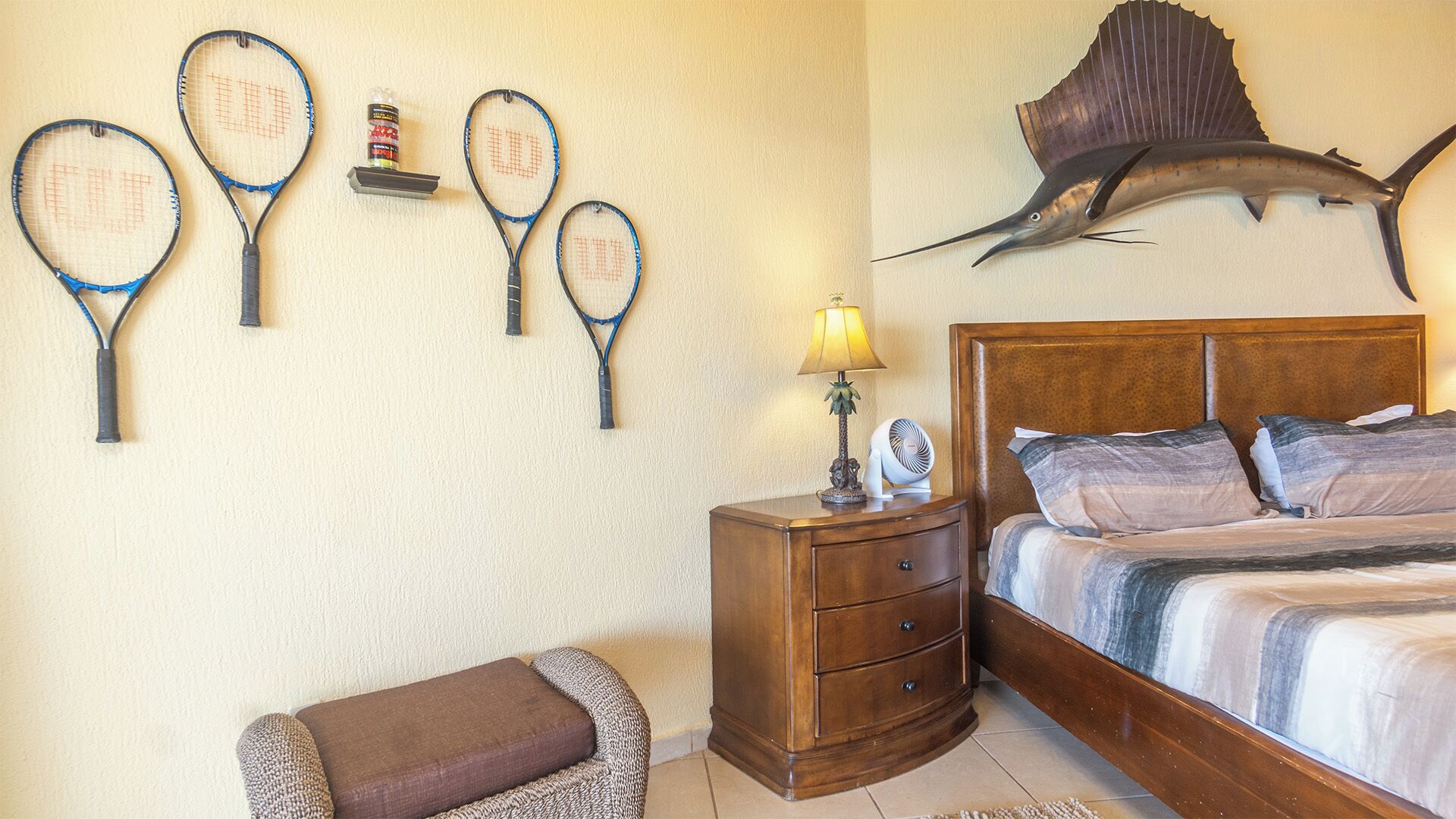 Une chambre à coucher comprenant un lit, une table de chevet, une lampe et des éléments de décoration murale, notamment un motif de poisson et des raquettes de tennis.
