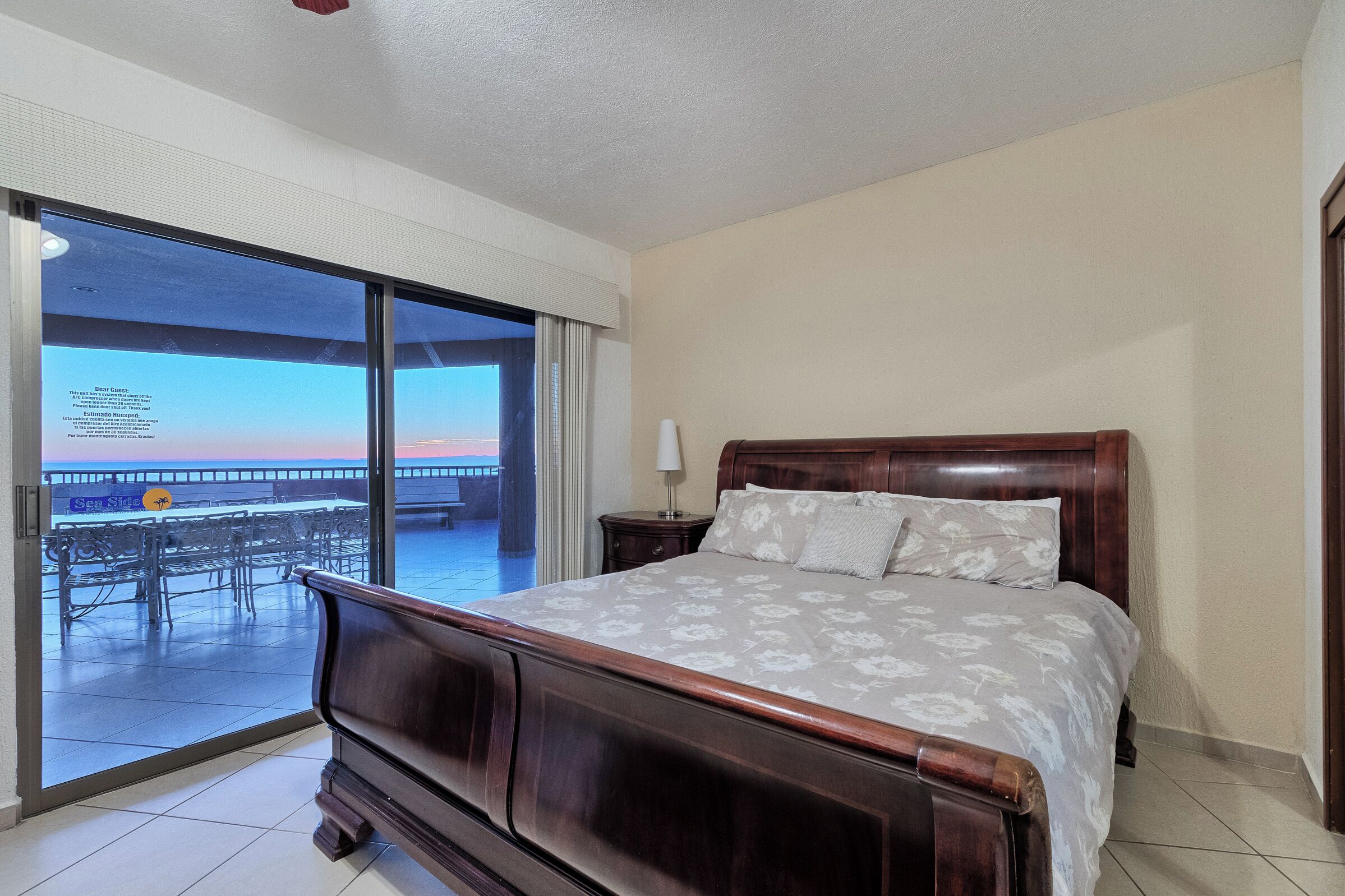A bedroom with a wooden bed, a nightstand, and a view of a beach and pier through a glass door.