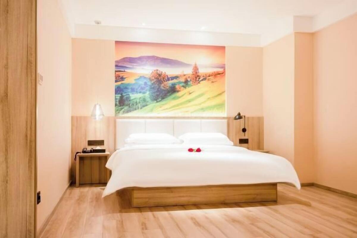 Une chambre à coucher comprenant un lit, une table de chevet, une lampe fixée au mur et un grand tableau représentant un paysage.