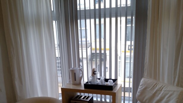 Ein Hotelzimmer mit einem Bett, einem weißen Sessel, einem kleinen Tisch mit einem Radio und einer Tasse sowie einem Fenster mit vertikalen Jalousien.
