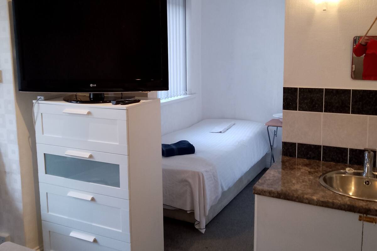 A compact hotel room with a bed, a TV, a small kitchenette, and a window with blinds.