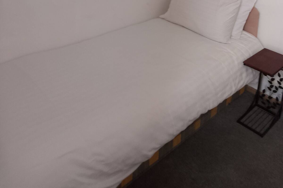 A single bed with a white mattress and pillow, a small bedside table with a book, and a wall-mounted picture.