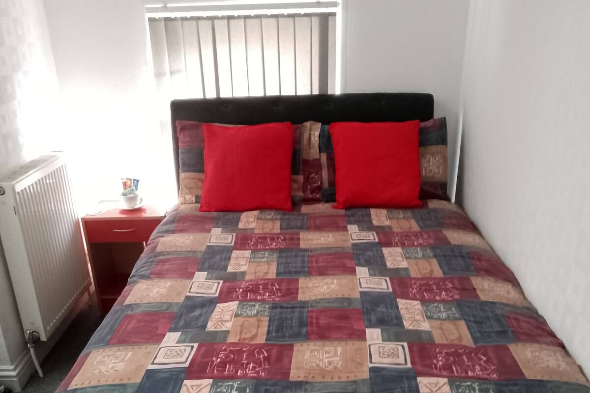 A bed with a quilted cover, a bedside table with a cup, and a window with blinds.