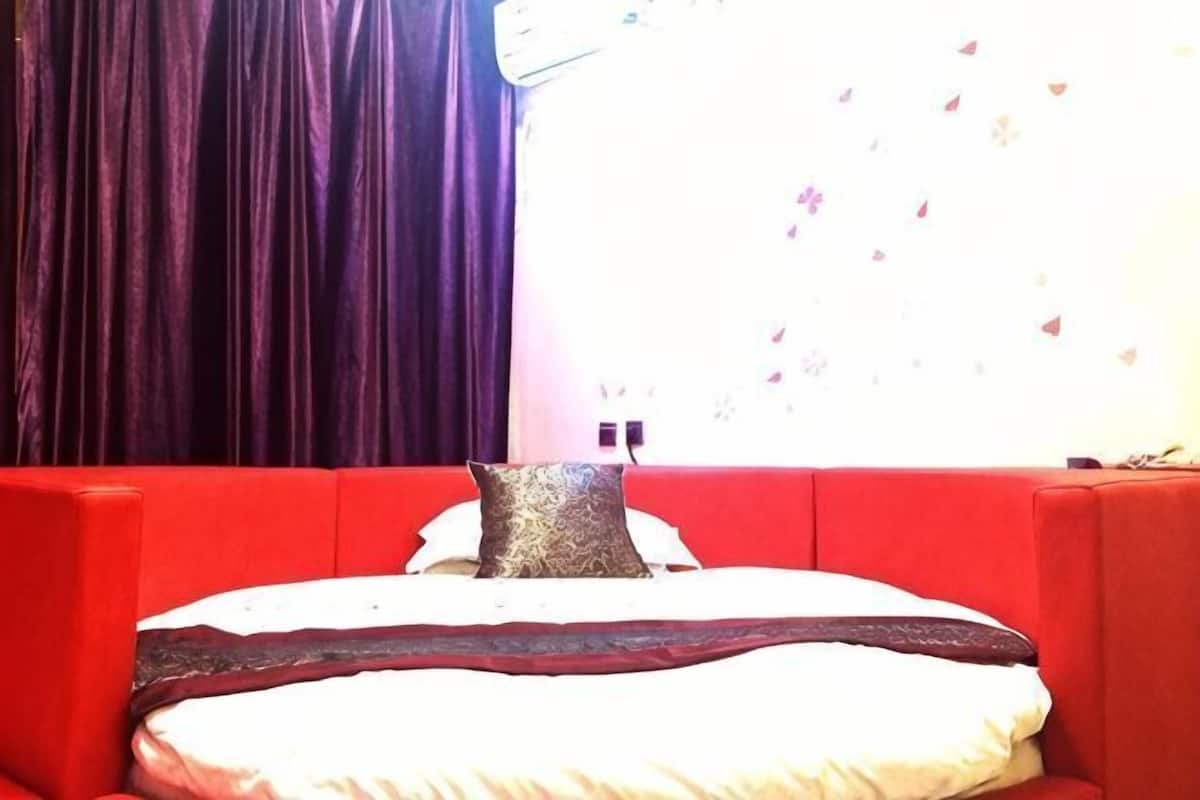 Un canapé-lit rouge avec un coussin à motifs, une fenêtre donnant sur un immeuble et un rideau.