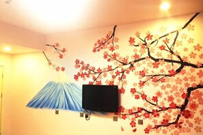 Quarto com televisão na parede, ventoinha no teto e parede decorada com motivos de flor de cerejeira.