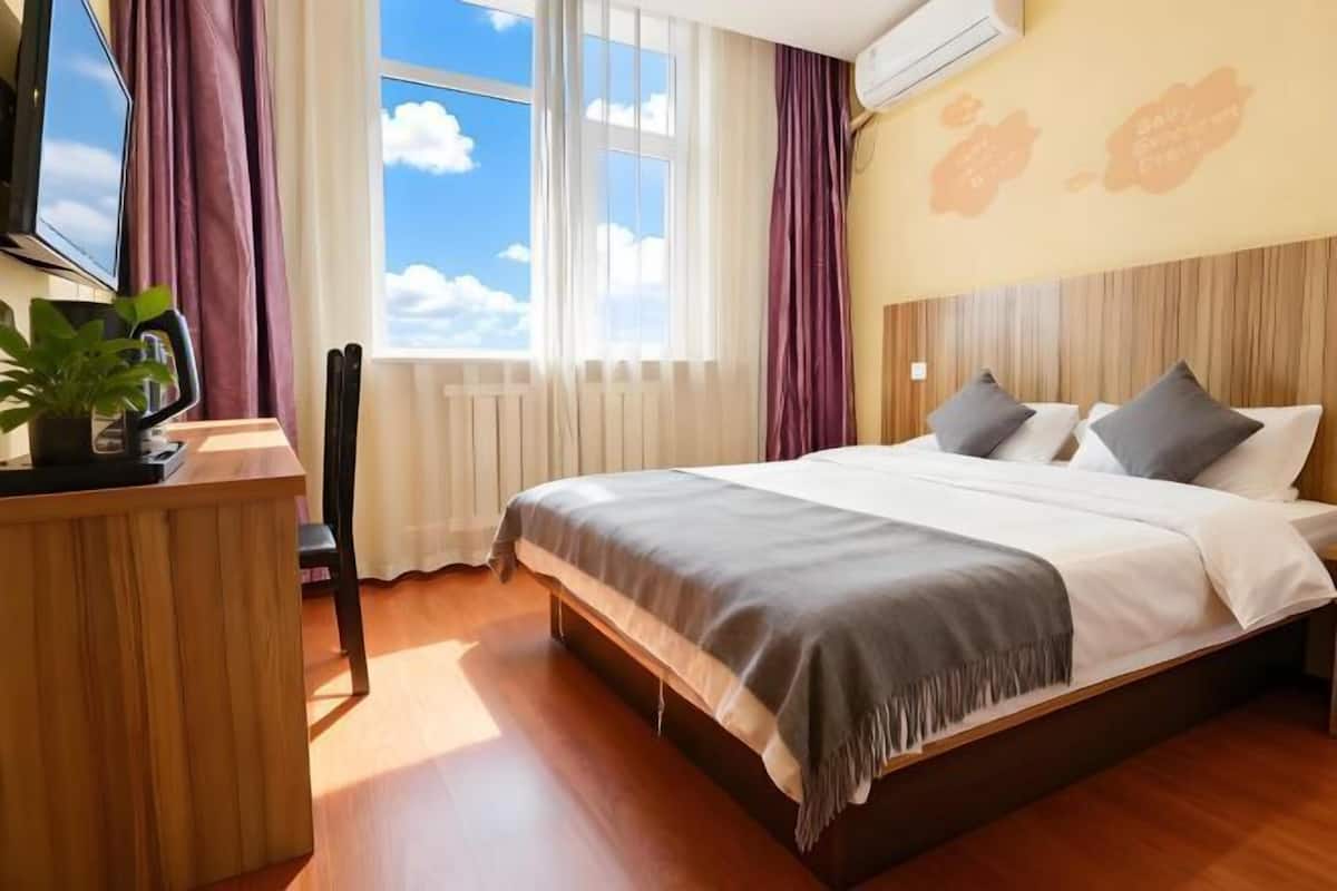 Une chambre d’hôtel avec un lit, un bureau, une chaise, une plante en pot et une fenêtre avec des rideaux.