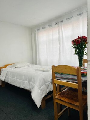 A single bed with white bedding, a wooden chair, a small table with a colorful cloth, and a vase with red flowers. - Enede Hostel Cusco (Cusco)