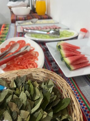 A table with various food items including sliced tomatoes, watermelon, and a basket of green leaves. - Enede Hostel Cusco (Cusco)