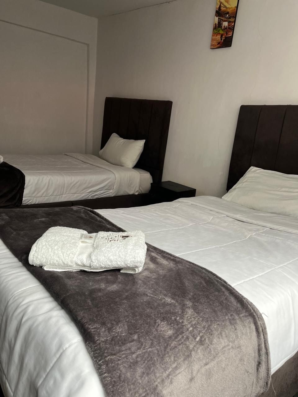 A hotel room with two beds, each with a dark headboard and white bedding. A folded towel is placed on the bed in the foreground.