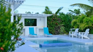 A swimming pool with blue-tinted water, surrounded by white lounge chairs and a small white structure with a screen. - Lois Estates: 1 bedroom home (Long Bay Caicos Islands)