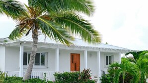 A white single-story house with a wooden door, surrounded by lush greenery and palm trees. - Lois Estates: 1 bedroom home (Long Bay Caicos Islands)
