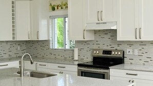 A modern kitchen with white cabinets, a stainless steel sink, a black stove, and a window with blinds. - Lois Estates: 1 bedroom home (Long Bay Caicos Islands)