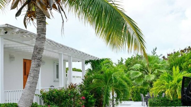 A white house with a covered porch, surrounded by lush tropical vegetation. - Lois Estates: 1 bedroom home (Long Bay Caicos Islands)