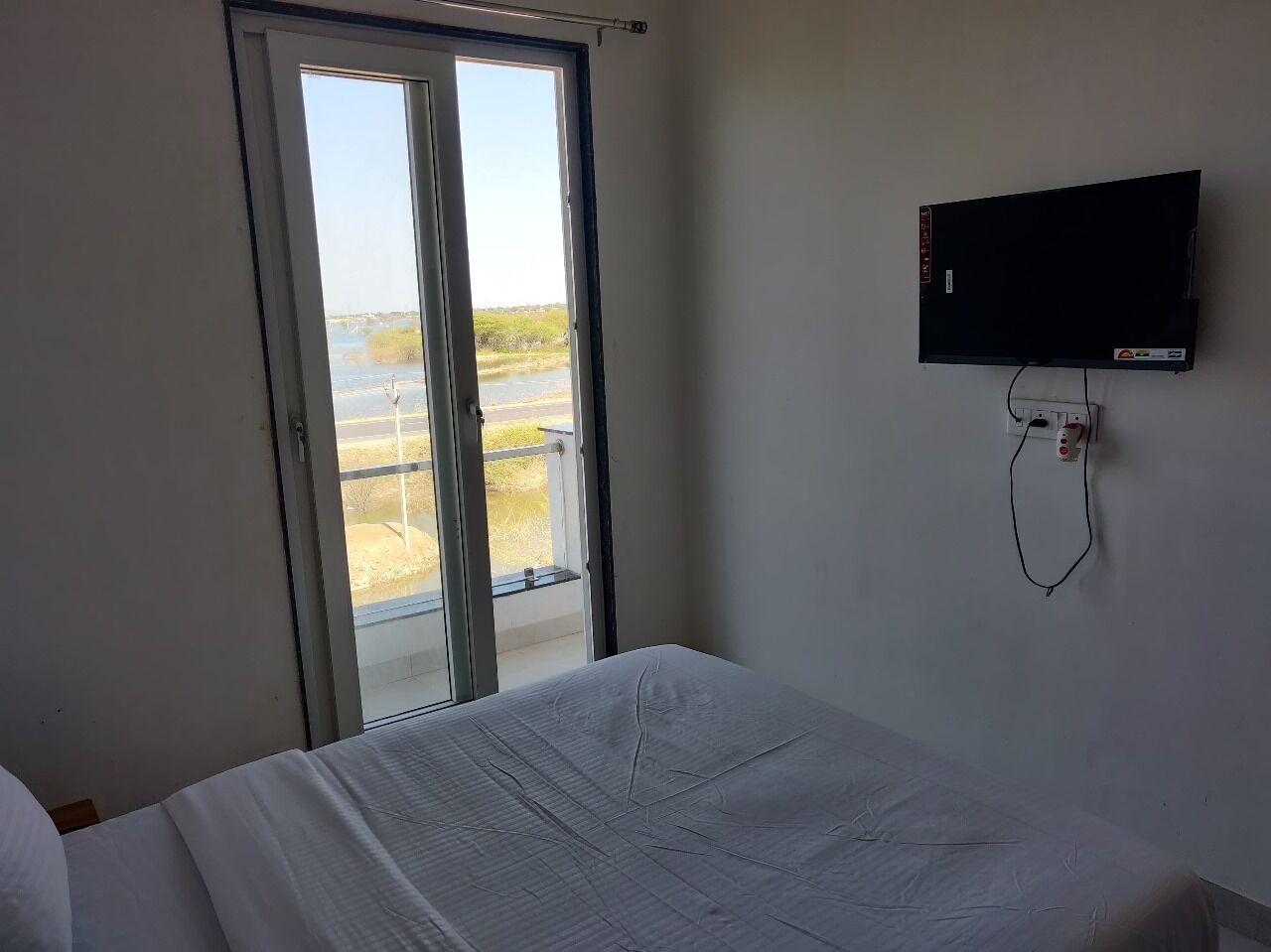 A hotel room with a bed, a TV mounted on the wall, and a sliding glass door leading to a balcony with a view of the beach.