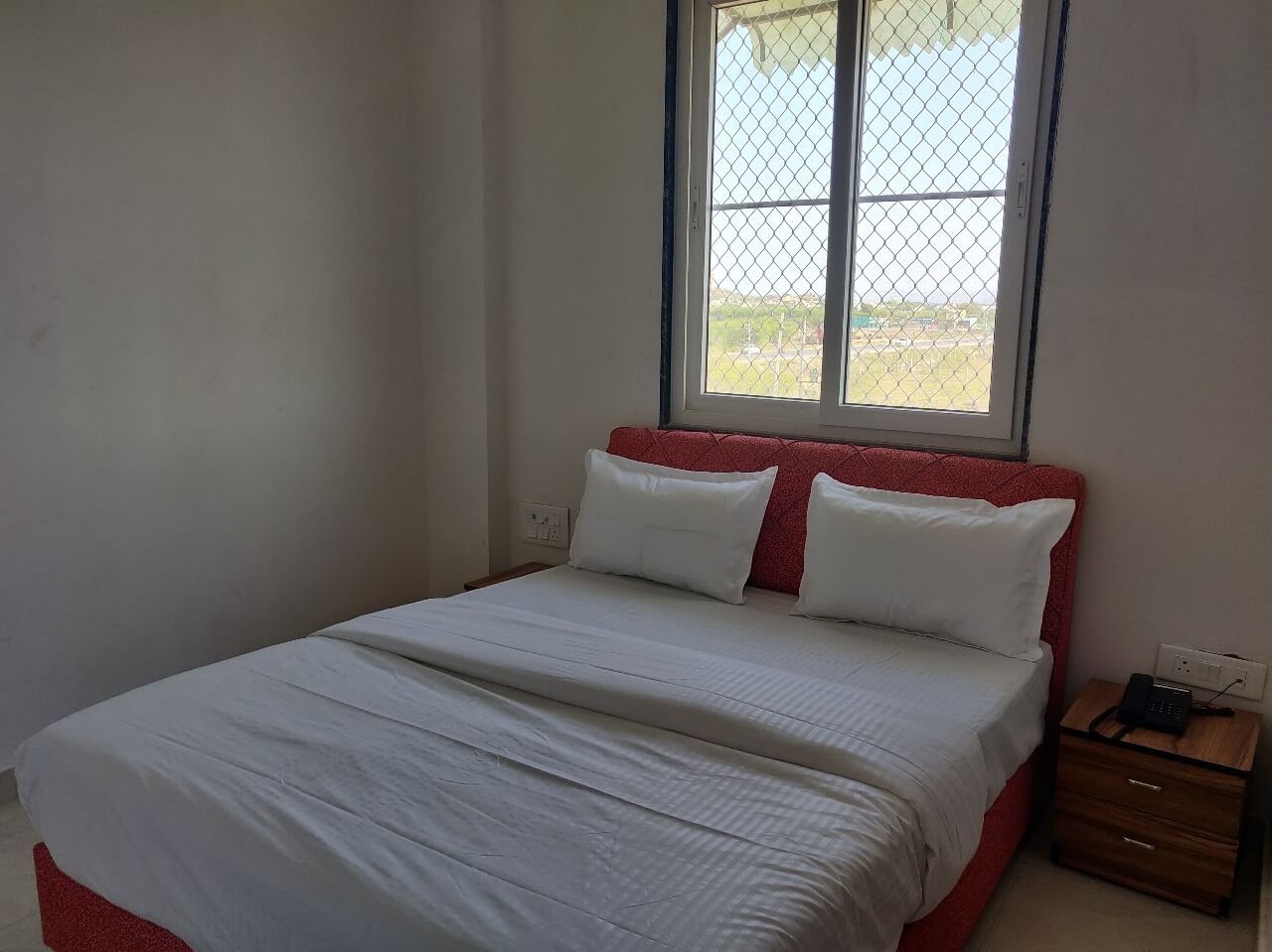 Een eenpersoonskamer met een rood hoofdeinde, witte beddengoed, een nachtkastje met een telefoon, en een raam met uitzicht.