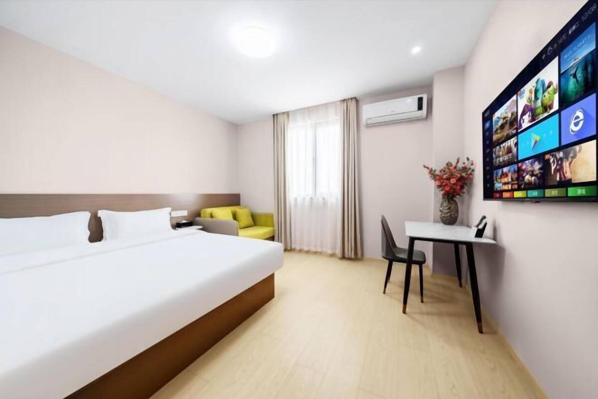 A hotel room with a bed, a small table with a chair, a TV mounted on the wall displaying various channels, and a window with curtains.
