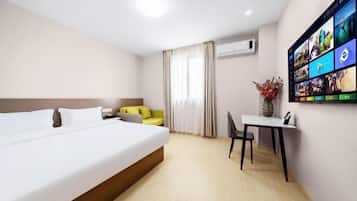 A hotel room with a bed, a small table with a chair, a TV mounted on the wall displaying various channels, and a window with curtains.