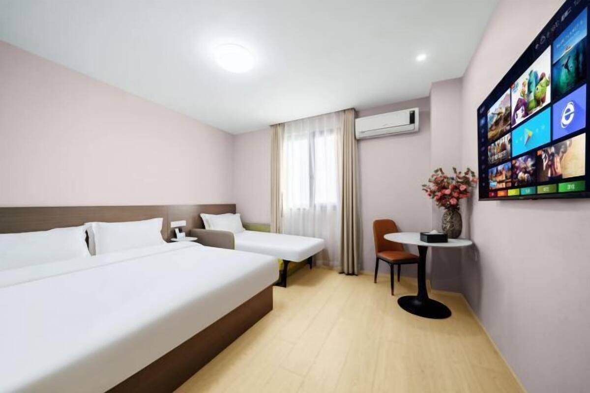 A hotel room with two beds, a small table with a vase, and a wall-mounted screen displaying various images.