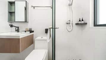 A modern bathroom with a glass shower enclosure, a white toilet, a wooden vanity, and a small shelf with toiletries.