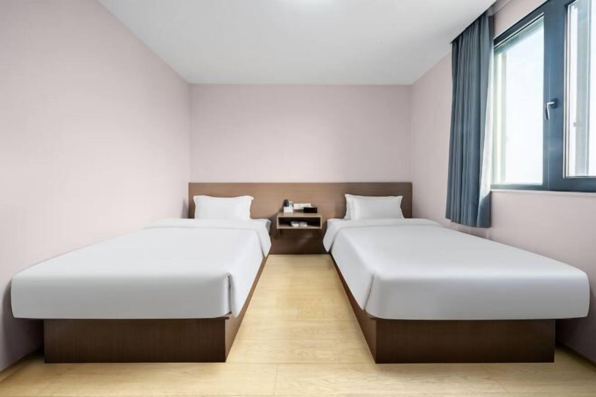 A hotel room with two single beds, a wooden headboard, a nightstand, and a window with curtains.