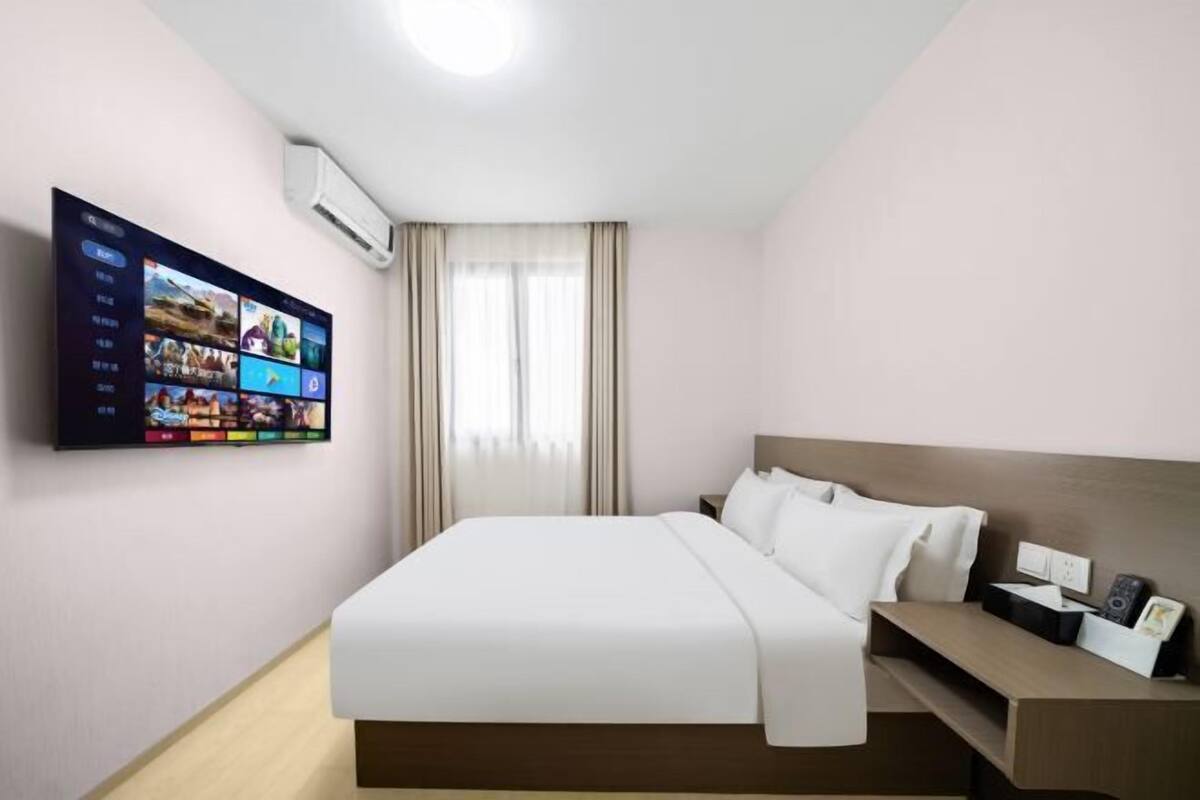 A hotel room with a bed, a bedside table, a TV mounted on the wall, and a window with curtains.