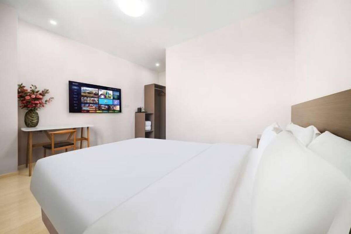 A hotel room with a bed, a desk, a chair, a TV on the wall, and a vase with flowers.