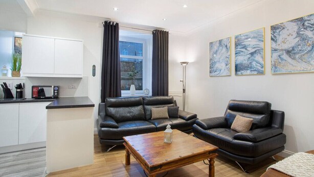 A modern living room with a black leather sofa set, a wooden coffee table, and a kitchen area with white cabinets and a microwave. - Selkies Retreat | Grampian Lettings Ltd (Aberdeen)