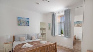 A bedroom with a wooden bed, a bedside table, a nightstand, a window with curtains, a view of the beach, and a painting on the wall. - Selkies Retreat | Grampian Lettings Ltd (Aberdeen)